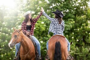 Due cowgirl amici con cavalli di fronte il di fronte direzione dare un' alto cinque foto