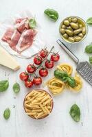 italiano o mediterraneo cibo cucina e ingredienti su bianca calcestruzzo tavolo. tagliatelle pene pasta olive oliva olio pomodori parmigiano formaggio prosciutto e basilico le foglie su bianca calcestruzzo tavola foto