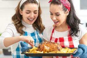 Due giovane casalinghe nel scacchi grembiuli dai un'occhiata e gusto appena fatto arrostito pollo e patate nel il cucina foto