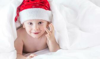 carino bambino ragazzo con Santa cappello su letto sotto bianca piumone foto