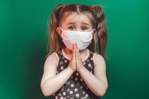 la bambina malata caucasica in maschera medica durante l'epidemia di coronavirus prega su sfondo verde primo piano