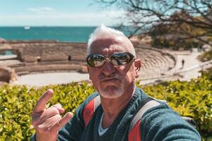 bello mezzo anziano uomo visitare taraco archeologico complesso, tarragona - contento turista assunzione un' autoscatto nel davanti di romano rovine foto