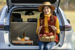 contento asiatico contadino ragazza trasporto produrre raccogliere con homegrown organici mela, schiacciare e zucca seduta su il auto tronco a Locale azienda agricola mercato durante autunno stagione per agricoltura Prodotto concetto foto