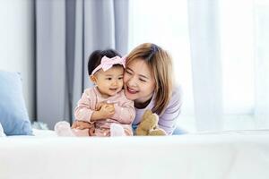 asiatico madre è Tenere sua bella sorridente bambino figlia mentre la spesa qualità tempo nel il letto per famiglia felicità e genitorialità concetto foto