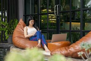 giovane attraente asiatico donna assunzione Nota e Lavorando al di sopra di il computer portatile nel riposo zona a il Hotel durante viaggio su vacanza, concetto di digitale nomade, viaggio liberamente, lavorando remoto su Internet in linea foto