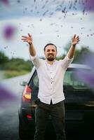 giovane sorridente uomo lanci su fiori di lupini vicino il macchina. tramonto o Alba, luminosa sera leggero foto