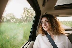 allegro positivo Riccio giovane donna nel casuale indossare seduta nel automobile sedile posteriore con fissato cintura di sicurezza e guardare lontano. foto