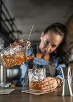 bella vista cameriera si concentra su scrosciante vecchio stile Bourbon cocktail in un ornamentale bicchiere nel un' pub foto