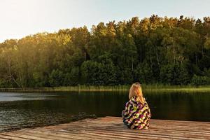 la bambina si siede su un molo di legno sul lago al tramonto in estate foto