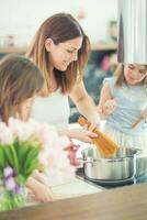 mamma con Due giovane Gemelli figlie nel il cucina cucinando spaghetti foto