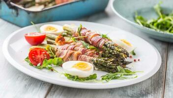 fresco asparago avvolto nel Bacon su un' bianca piatto con rucola pomodori e uova foto