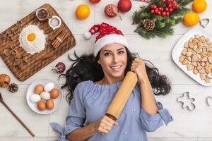 yound e contento donna strettamente Tenere un' rullo mentre posa su il terra con tematico cose piace. Natale ingredienti, Farina e uovo, arance, cottura al forno le forme e rosso Santa cappello foto