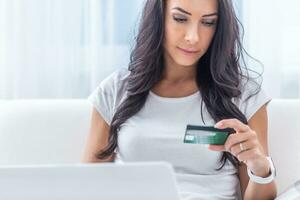 brunetta giovane donna guardare a il credito carta fare in linea pagamento nel il bianca ambiente di sua Casa foto