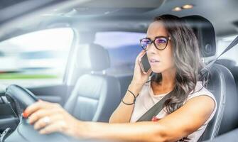 donna nel bicchieri guida un' auto chiamate su un' cellula Telefono, Tenere timone ruota con solo uno mano foto