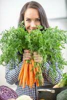 giocoso giovane donna nel sua cucina Tenere fresco carota nel tutti e due mani - dieta verdura e brughiera concetto foto