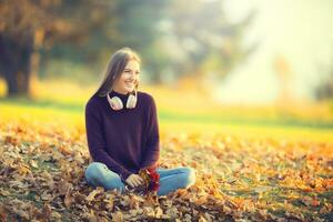 ritratto di contento smilling giovane ragazza con cuffie nel autunno parco foto