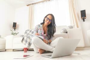 bellissimo donna nel bicchieri sorridente con un' inclinata testa mentre seduta su il vivente camera pavimento Lavorando su un' pc e bloc notes foto