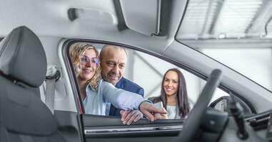 moglie Spettacoli sua marito Caratteristiche di un' nuovo auto nel il concessionaria, sorridente, mentre il i saldi assistente sta a parte foto