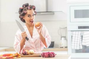 bene guardare donna sta nel il cucina Tenere coltello e un cipolla, indossare capelli bigodini e notte abito. foto