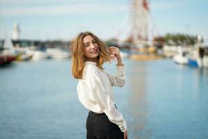 Visualizza a partire dal Indietro. giovane donna trasformato in giro e guardare dritto a telecamera. allegro bella ragazza in posa contro porto marittimo. ragazza indossare casuale Abiti ang sorridente. a piedi di mare su soleggiato giorno. foto