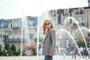 donna d'affari a Fontana. sofisticato donna con occhiali da sole. ragazza a piedi in giro città. sfondo di bellissimo architettonico edifici. donna indossare giacca e jeans. sfocato sfondo. foto