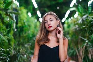 donna nel giardino con tropicale impianti. ritratto di bellissimo ragazza con lungo capelli in posa per telecamera. giovane donna indossare nero vestito su magro cinghie sta tra verde impianti dentro preparativi foto