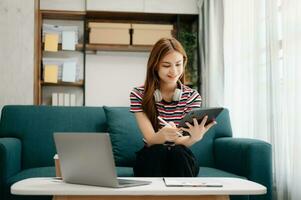 femmina studenti Nota a partire dal il libri a il asiatico ragazza biblioteca seduta a divano utilizzando il computer portatile computer e tavoletta per ricerca un in linea informazioni. foto