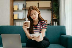 femmina studenti Nota a partire dal il libri a il asiatico ragazza biblioteca seduta a divano utilizzando il computer portatile computer e tavoletta per ricerca un in linea informazioni. foto