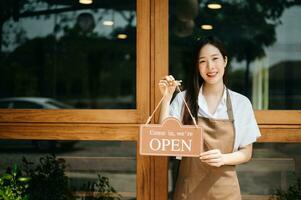 giovane femmina imprenditore sospeso un' benvenuto cartello nel davanti di un' caffè negozio. bellissimo cameriera o hostess Tenere un' tavoletta preparazione foto