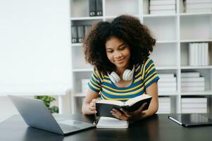 femmina studenti Nota a partire dal il libri a il africano ragazza biblioteca seduta a il scrivania utilizzando il computer portatile computer e tavoletta per ricerca un in linea informazioni. foto