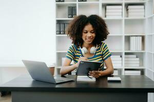 femmina studenti Nota a partire dal il libri a il africano ragazza biblioteca seduta a il scrivania utilizzando il computer portatile computer e tavoletta per ricerca un in linea informazioni. foto