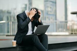 frustrato giovane asiatico uomo d'affari Lavorando su un' il computer portatile computer seduta a il suo Lavorando posto nel al di fuori ufficio foto