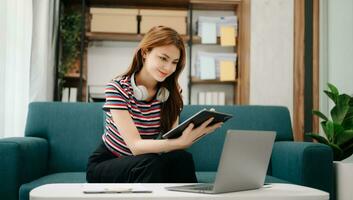 femmina studenti Nota a partire dal il libri a il asiatico ragazza biblioteca seduta a divano utilizzando il computer portatile computer e tavoletta per ricerca un in linea informazioni. foto