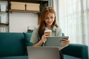 femmina studenti Nota a partire dal il libri a il asiatico ragazza biblioteca seduta a divano utilizzando il computer portatile computer e tavoletta per ricerca un in linea informazioni. foto