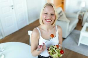 ritratto di un' contento giocoso ragazza mangiare fresco insalata a partire dal un' ciotola nel sua cucina. bellissimo in forma donna mangiare salutare insalata dopo fitness allenarsi foto