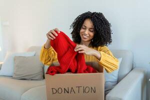 contento africano giovane donna sedersi su divano incollato Abiti nel donazione scatola a casa, cura birazziale femmina volontario mettere abbigliamento nel scatola di cartone pacchetto, donare per bisognoso le persone, riutilizzare, riciclare concetto foto