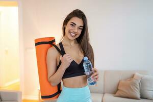 bellissimo giovane donna nel gli sport indossare è Tenere un' yoga stuoia e un' bottiglia di acqua, guardare a telecamera e sorridente foto