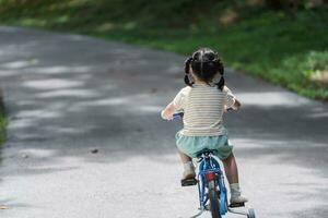 indietro lato Visualizza di asiatico bambino ragazza bambino apprendimento per cavalcata bicicletta nel il parco giardino. formazione scolastica concetto per ragazzo pratica Ciclismo a parco, bambino sport concetto. foto