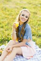 giovane contento donna detiene un' bicchiere di arancia succo nel sua mano mentre seduta su un' picnic coperta foto