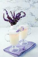 caffè con latte, schiuma e lavanda nel bicchieri e un' mazzo di fiori nel un' brocca su un' bianca tavolo. verticale Visualizza foto