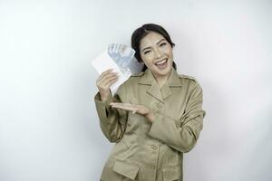 un' sorridente giovane governo lavoratore donna è indossare cachi uniforme Tenere denaro contante i soldi nel indonesiano rupia isolato di bianca sfondo foto