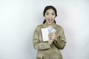 un' sorridente giovane governo lavoratore donna è indossare cachi uniforme Tenere denaro contante i soldi nel indonesiano rupia isolato di bianca sfondo foto