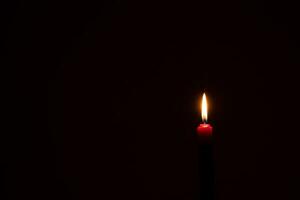 singolo ardente candela fiamma o leggero raggiante su un' piccolo rosso candela su nero o buio sfondo su tavolo nel Chiesa per Natale, funerale o memoriale servizio con copia spazio. foto