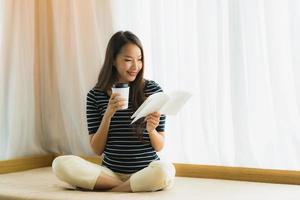 Ritratto bella giovane donna asiatica libro di lettura e tenere la tazza di caffè o tazza sul divano in soggiorno area living foto