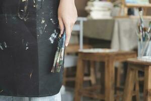 un' avvicinamento di il artista mani indossare un grembiule spalmato con dipingere. stringendo molti spazzole e pennelli. foto