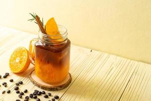 un bicchiere di caffè nero americano ghiacciato e uno strato di succo di arancia e limone decorato con rosmarino e cannella su uno sfondo di legno foto