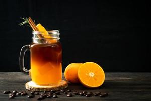un bicchiere di caffè nero americano ghiacciato e uno strato di succo di arancia e limone decorato con rosmarino e cannella su uno sfondo di legno foto