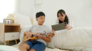 un' ragazza dire bugie nel letto giocando tavoletta e un' ragazzo seduta accanto letto giocando chitarra nel un' Camera da letto. foto