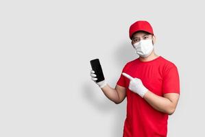 impiegato dell'addetto alle consegne con berretto rosso maglietta bianca uniforme maschera per il viso con applicazione di telefono cellulare nera isolata su sfondo bianco foto