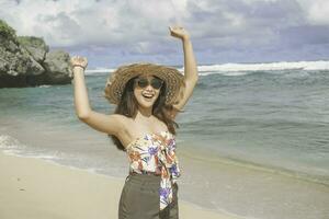 un' contento giovane asiatico donna indossare spiaggia cappello è in posa per il telecamera con sua mani aprire, esprimendo la libertà e spensierato vacanza a il spiaggia nel gunungkidul, Indonesia foto